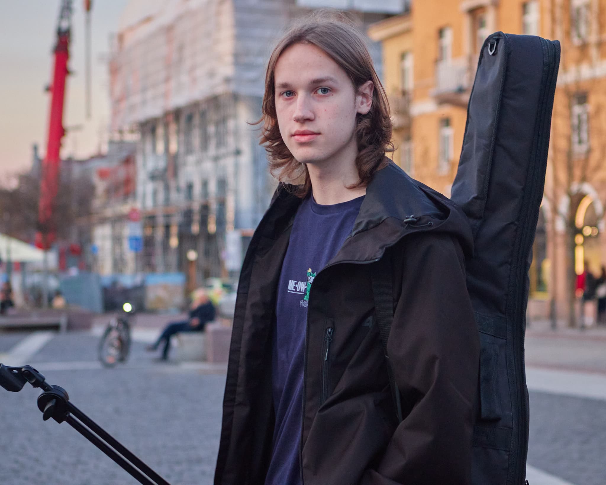 Jaunas vyras su gitaros dėklu pozuoja miesto fone