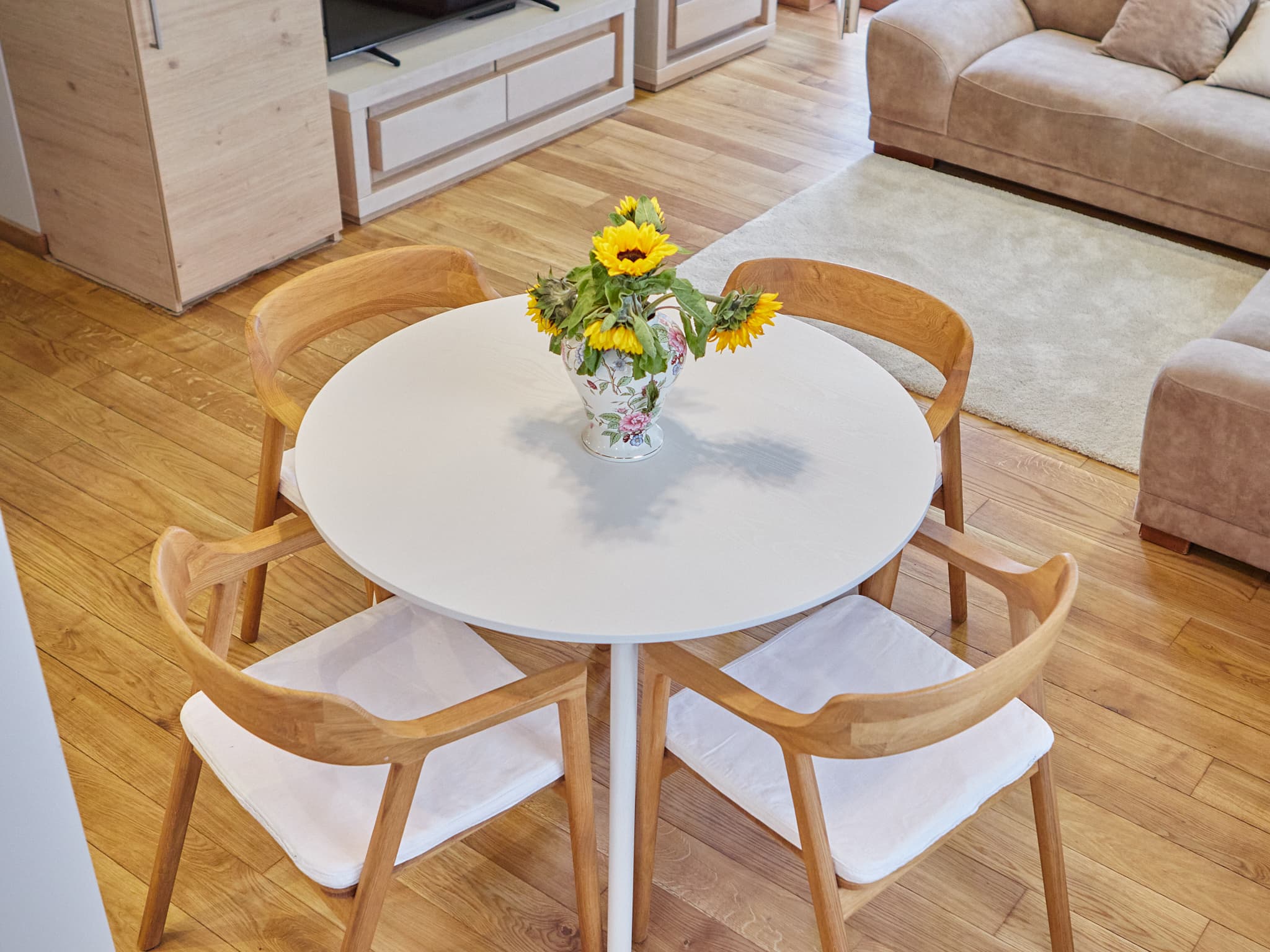 Apvalus stalas su gėlėmis svetainės erdvėje, matomas televizorius ir sofa