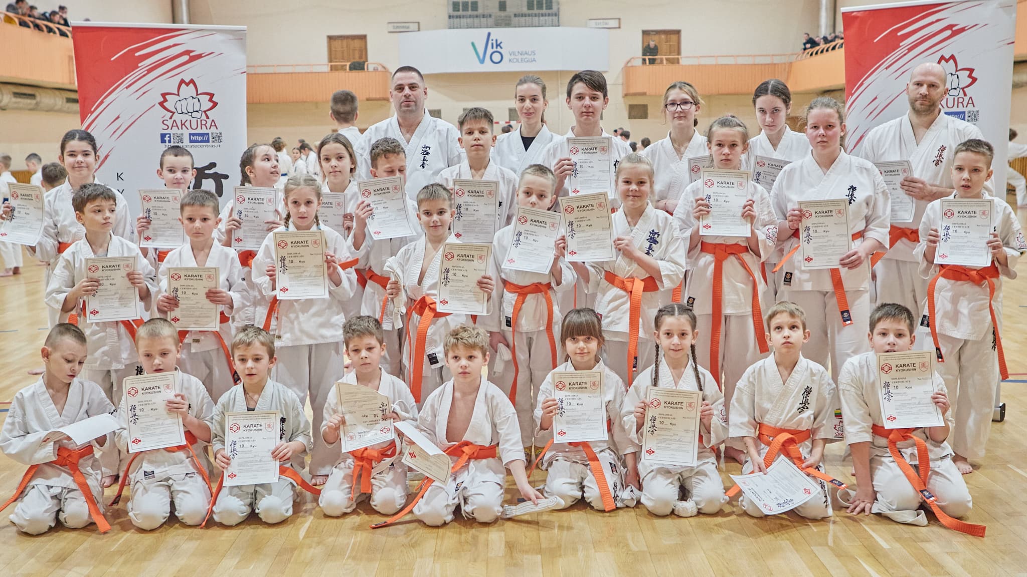 Vaikai ir treneriai su diplomais pozuoja sporto salėje