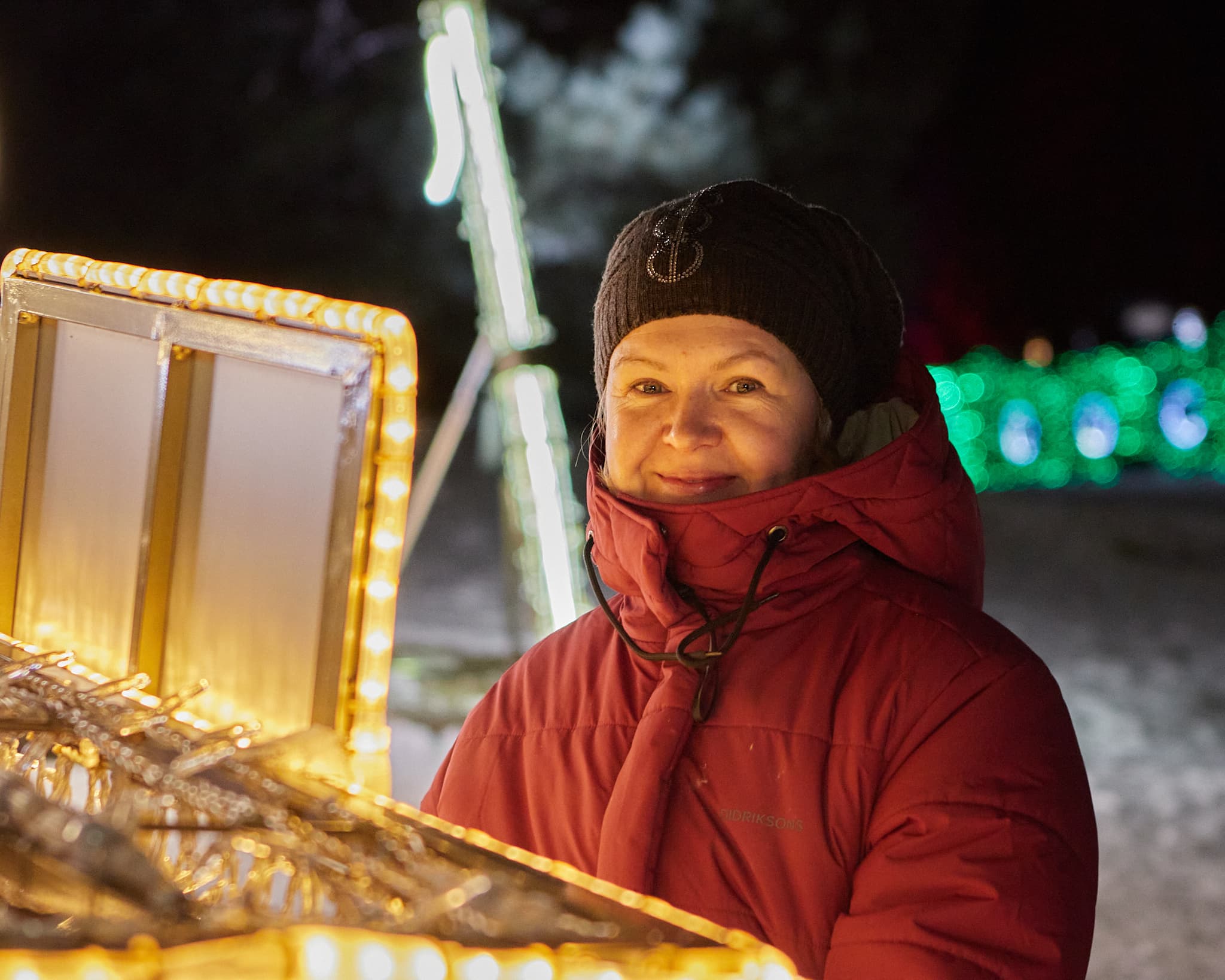 Moteris su raudona striuke pozuoja prie šviečiančios dekoracijos žiemą